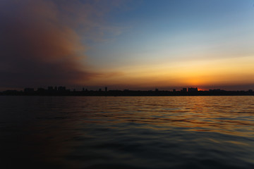 Obraz premium Beautiful sea landscape. Bright warm sunset in the sea near the coastline with the city's silhouettes. Sun hides behind the house. Colors of the sky are reflected in the water