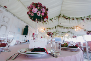 Beautiful decoration of the wedding banquet under the awning in pink, burgundy and white tones. Table setting in a hall with plates, cutlery, glasses, napkins and cards, decorated floral compositions