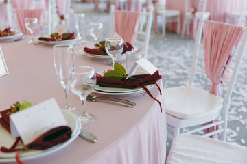 Beautiful decoration of the wedding banquet under the awning in pink, burgundy and white tones. Table setting in a hall with plates, cutlery, glasses, napkins and cards, decorated floral compositions