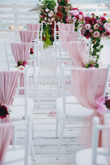 Beautiful decoration of the wedding ceremony in pink, burgundy and white tones on wooden pier. Chairs decorated with a thin cloth, beads and floral compositions of roses and peonies