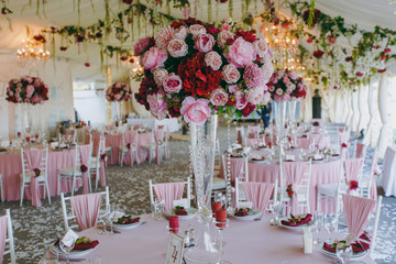 Beautiful decoration of the wedding banquet under the awning in pink, burgundy and white tones. Hall with dining tables and chairs decorated with thin cloth, bouquets, flower garlands and confetti