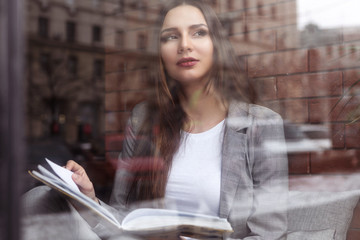 Brunette caucasian young woman sitting near the window on the brick wall and wooden surface background in grey suit and white blouse and reading a book and dreaming making plans