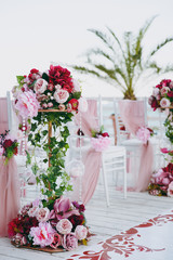 Beautiful decoration of the wedding ceremony in pink, burgundy and white tones on wooden pier. Chairs decorated with a thin cloth, beads and floral compositions of roses and peonies