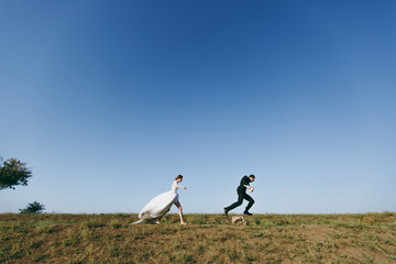 Obraz premium Beautiful wedding photosession. Young charming bride in white lace dress with long plume runs for her fiancé in a black suit on walk around the big green field against blue sky background