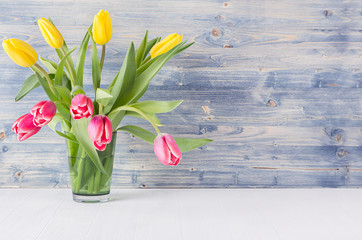 Bouquet red and yellow tulips in green glass vase on blue shabby wooden background. Festive easter springtime interior.