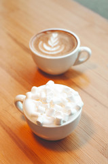 Two cups of cappuccino with latte art and marshmallow on wooden background. Beautiful foam, greenery ceramic cups, stylish toning, place for text.