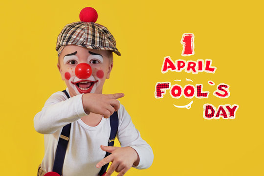 Happy clown show finger text 1 april fools day. Funny little child boy in costume, makeup, red nose cheerful clown, bright yellow background. Emotional mischievous portrait kid actor.copy space.