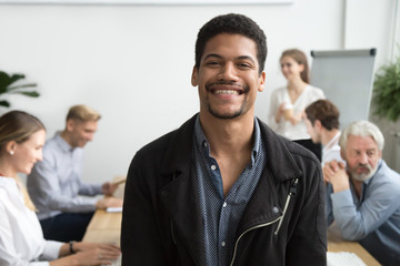 Smiling african american office employee or leader looking at camera with diverse colleagues at background, happy young black manager, professional coach or team member posing, head shot portrait