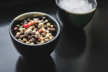 Close up view of coarse salt and allspice, white, green, black and white pepper in ceramic bowls on black background. Vintage toned. Spices background