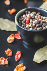 Close up view of coarse salt and colorful allspice pepper in ceramic bowls with bay leaves and red dried chilly peppers on black background. Spices background