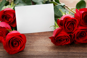 Bouquet of red roses with sheet of blank paper on brown wooden table