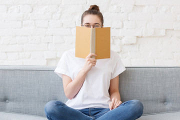 Young hipster teen with bun in round eyeglasses and white t-shirt is hiding behind old book