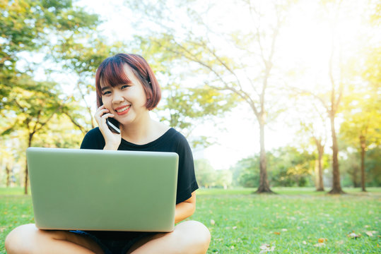 Cute asian woman smiles and talking on mobile phone while sitting in park spring day. Asian woman using on smart phone and laptop with feeling relax and smiley face. Lifestyle and technology concepts.