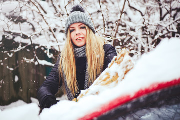 Winter portrait of a young woman cleaning snow from a car. Beauty blonde Model Girl laughs and cheerfully cleans the snow. Beautiful young woman outdoors