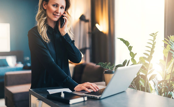 Young smiling businesswoman in black blouse is standing indoor, working on computer, while talking on cell phone. Girl freelancer, entrepreneur works at home.Online marketing, education, distance work