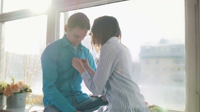 Sunlit young couple sitting by the window and kissing, both in shirts, the girl correcting the guy's tie