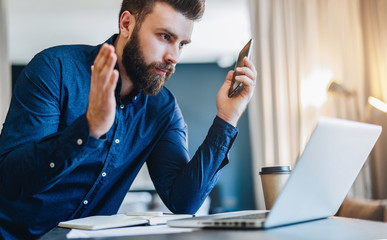 Young bearded businessman sits in front of computer and looks at laptop screen with astonishment, raising his hands up. Guy is surprised by information received, an unexpected e-mail. Distance work.