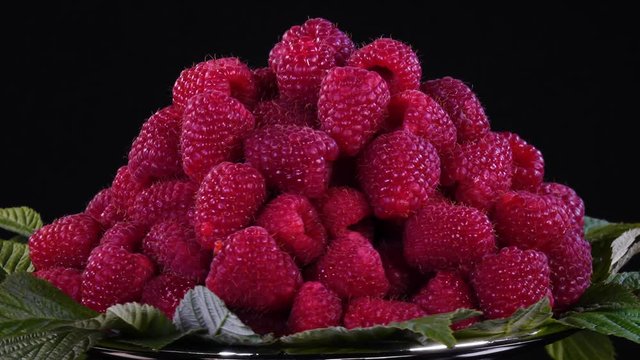 Fresh raspberries with green leaves rotate on silver cake stand against black background. 4K. Closeup view of sweet fruit.
