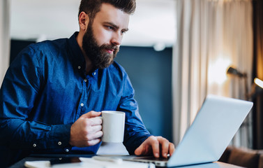 Young serious bearded businessman working on computer at table, drinking coffee. Man analyzes information, data, develops business plan. Freelancer working. Online marketing, education, e-learning.