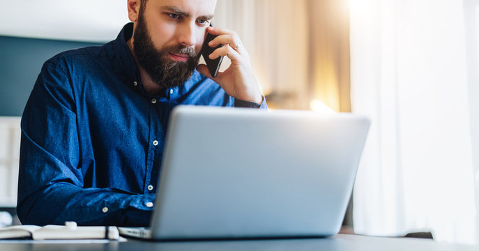 Front view. Young bearded businessman is sitting at table in front of computer, talking on cell phone. Freelancer, entrepreneur works at home. Telephone conversations. Distance work, online education.