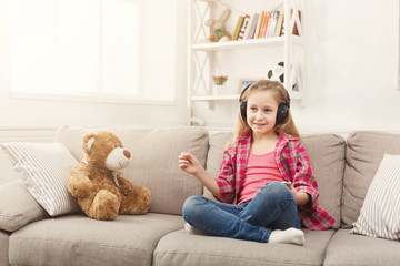 Happy little female child and her teddy bear listening to music on sofa at home