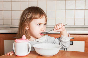 Little gloomy girl eats and drinks at the table in the kitchen