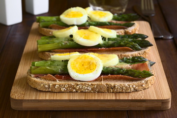 Baked green asparagus, ham, hard-boiled egg and grated cheese warm sandwich on wooden board, photographed on dark wood with natural light (Selective Focus, Focus on the front of the first sandwich)