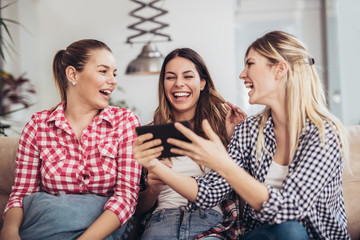 Obraz premium Three best friends using digital tablet together. Women sitting on sofa having fun surfing on the internet using smart digital pc tablet modern device