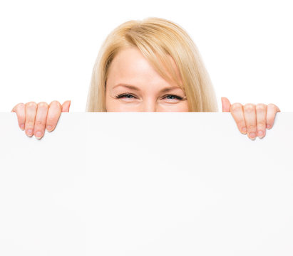 Young woman with surprised eyes peeking out from behind billboard paper poster. Businesswoman holding big white banner, isolated on white background.