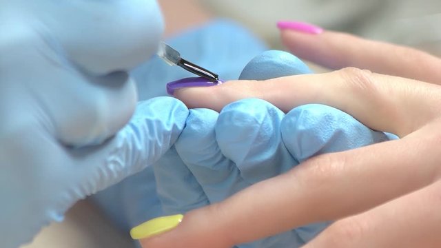 Manicurist covers nail with fixer close up. Nail specialist in blue gloves fixing polished nail to client with transparent fixer, slow motion.