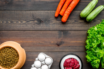 Dry pet food with natural ingredients. Raw meat, vegetables zucchini and carrot near eggs on dark wooden backgroud top view copy space