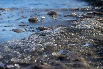 Close up picture of beach's surface. Ice is melted. Sand is wet and dirty. Some sun lights reflects on melted surface of ice cover.