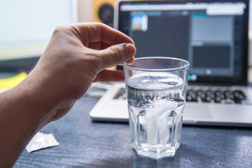 hand through pill of aspirin in glass of water. laptop on background. overloaded with office work