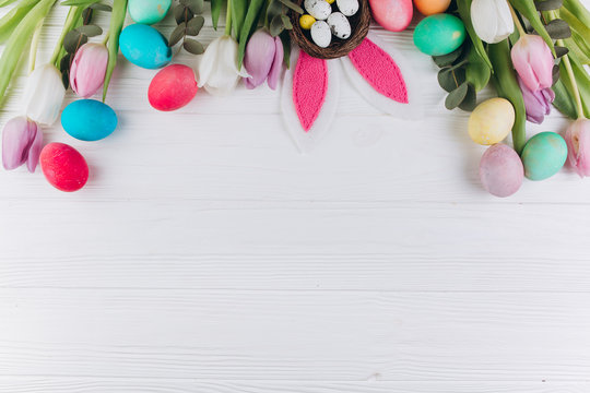 Easter composition with colored eggs, rabbit ears, nest and tulips on a white wooden background.