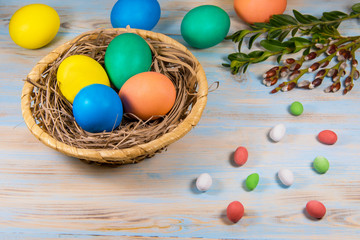 Fototapeta na wymiar Easter eggs in a wooden basket, painted in different color on a blue background with a place for the inscription next to chokolate candy and green twigs