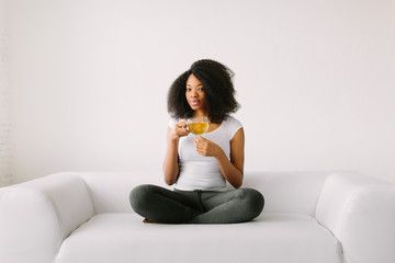 An African American young women sitting in the lotus position on white bed with cup of tea