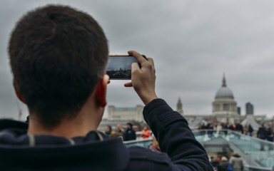 Young man taking a picture using his smartphone of the Millenium Bridge and St. Paul's Cathedral in London, England, UK.