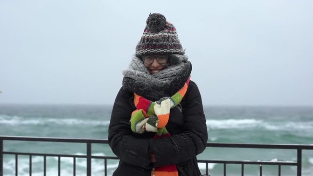 Portrait of attractive hipster woman 20s wearing glasses dressed warmly, while having day off by ocean in winter season under snowfall