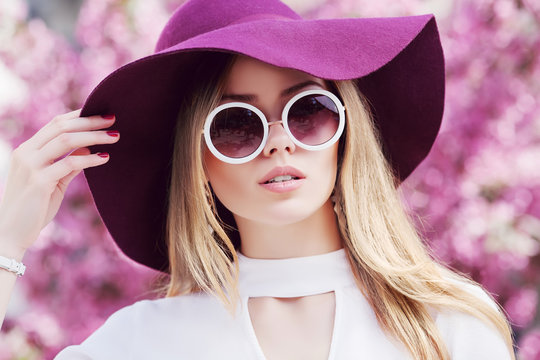 Outdoor close up portrait of young beautiful girl wearing stylish round sunglasses, trendy violet hat, white shirt, posing in street. Pink blooming trees on background. Spring fashion concept. 