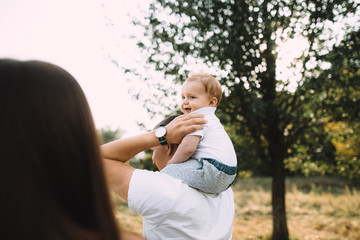 Happy spouses spend time together with their young son outside the city. The baby sits on shoulders of his father and laughs