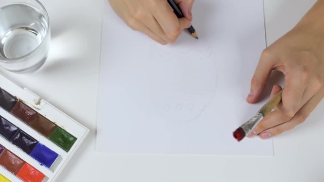 Unrecognizable woman draws a pencil sketch on paper Easter card. On the table is a palette with paints and a glass of water.