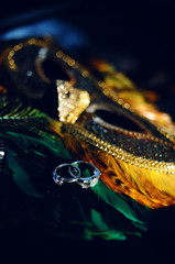 Wedding rings, jewelry and symbols attributes taken with selective focus on peacock colored carnival mask. Holiday, celebration. Macro. Blur. Bridal bouquet on background