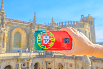 Tourism and travel concept in Portugal. Close up of mobile phone with Portugal flag cover taking photos of tourist place in Templar city. Convent of Christ or Templar fortress on blurred background.