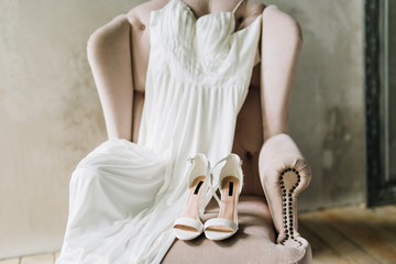 close-up of wedding shoes on a pink armchair next to a wedding dress against a gray concrete wall
