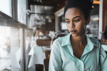 Portrait of a young beautiful Brazilian girl sitting in the cafe and wistfully looking aside; a charming African American female in a teal striped shirt is waiting for her friend in the restaurant