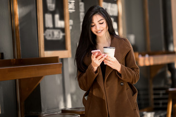 Beautiful girl uses a phone and drinks coffee, sitting in a cozy cafe.