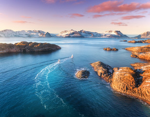 Aerial view of fishing boats, rocks in the blue sea, snowy mountains and colorful purple sky with red clouds at sunset in winter in Lofoten islands, Norway, Landscape with two ship. Top view. Travel - obrazy, fototapety, plakaty