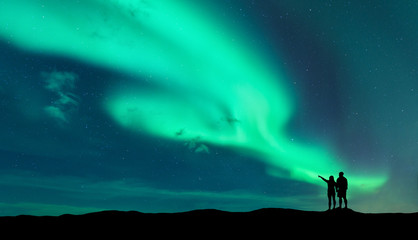 Aurora borealis und Silhouette eines stehenden Mannes und einer Frau, die mit dem Finger auf Nordlichter zeigen. Lofoten-Inseln, Norwegen. Aurora. Himmel mit Sternen und Polarlichtern. Nachtlandschaft mit Aurora und Paar