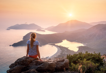Young woman sitting on the top of rock and looking at the seashore and mountains at colorful sunset...