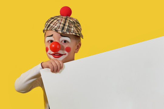 Funny child clown with blank white paper. Cute little boy clown in makeup,red nose. Little cheerful boy jester keep sheet with copy space for your text. Concept birthday, holiday,celebration, party.